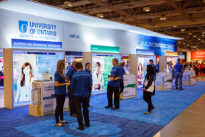 An image of one side of the new UOIT booth at the Ontario Universities Fair. A diverse group of staff are waiting beside counters for each faculty..