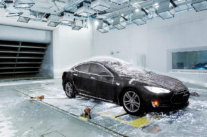 A black Tesla car in the ACE climatic wind tunnel, covered in ice and icicles, with a trail of smoke indicating the flow of wind over the vehicle.