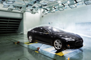 A black Tesla car in the ACE climatic wind tunnel, subjected to simulated rain, with a trail of smoke indicating the flow of wind over the vehicle.