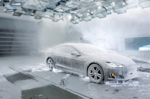 A black Tesla car in the ACE climatic wind tunnel, covered in snow and icicles, with clouds of mist and snow billowing around the vehicle.